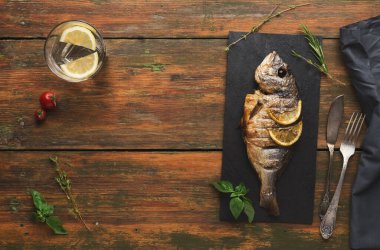 Bir yiyecek koleksiyonu ahşap bir masada düzenlenir. Izgara bir balık, limon dilimleriyle siyah bir taş tabağa konur. Yakınlarda bir bardak su var. Taze otlar etrafı sarmış..