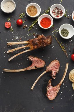 Kuzu pirzolası, çeşitli baharatlar, otlar ve taze malzemelerle birlikte koyu bir yüzeye yerleştirilir. Bu sahne, baharatlı bir yemeğin hazırlanmasını gösteriyor..