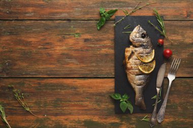 Izgara balık taze otlar ve limonla çevrili siyah servis tabağında yatar. Kırsal bir ahşap masa bu yemek hazırlığı için bir ortam görevi görüyor. Çatal ve bıçak çanağın yanına konur..