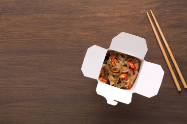 Paket servis kutusu ahşap bir masada duruyor. Kutunun içinde baharatla karıştırılmış erişteler var. Kutunun yanında kullanıma hazır çubuklar var. Yemek hızlı bir lokma için davet edici görünüyor..