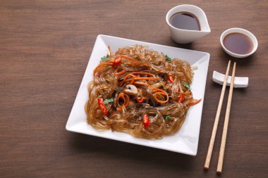 Bir tabak, içinde parlak renkler ve malzemeler olan bir erişte tabağı tutar. Yanında yemek çubuklarıyla birlikte soslu iki küçük kase var. Ahşap bir masa üzerinde ayarlar basittir..