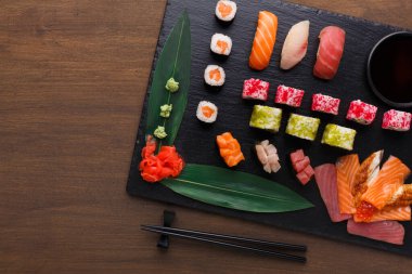 Suşi koleksiyonu siyah bir tabağın üzerinde duruyor. Farklı suşi türleri düzgün bir şekilde düzenlenir. Suşi rulo, sashimi ve renkli malzemeler içerir. Yeşil wasabi ve çubuklar mevcut..