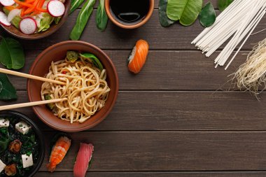 Farklı Asya yemekleri ahşap bir masada hazırlanır ve düzenlenir. Kaselerde erişte, salata, suşi ve sebze var. Taze malzemeler ilave renk ve çeşitlilik için yerleştirilir.