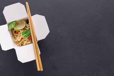 Farklı yemek türleri paket kutusuna konur. Limon ve otlar lezzet için üstte. Yemek çubukları yakınlarda. Sahne, yiyecekleri ön plana çıkaran karanlık bir yüzeye kurulmuştur..