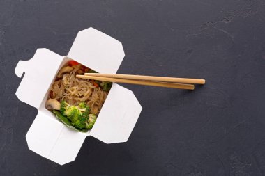 Paket servis kutusu açık ve içinde erişte ve sebze olduğunu gösteriyor. Yemeklerin yanındaki kutuda dinlenen yemek çubukları var. Arka plan karanlık ve basit, dikkat dağıtacak bir şey yok..