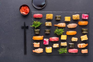 Siyah bir tabakta düzgünce düzenlenmiş bir suşi koleksiyonu. Çeşitli suşi türleri mevcuttur, farklı balık ve sarmalar da dahil. Soya sosu ve zencefil turşusu da dahil..