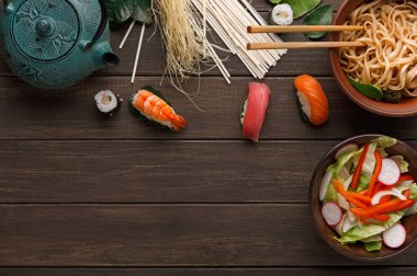 Tahta bir masanın üzerine yerleştirilmiş yiyecek koleksiyonu. Bulaşıklar arasında suşi, salata, erişte ve çaydanlık var. Çubuklar kaselerin yanına konur. Taze malzemeler basit bir düzende gösterilir.