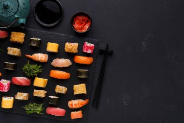 Bir kolaj siyah servis tahtasına düzgünce yerleştirilmiş çeşitli suşi türlerini gösterir. Tahtada renkli parçalar yer alıyor ve yanında da küçük kaseler dolusu soya sosu ve zencefil turşusu var..