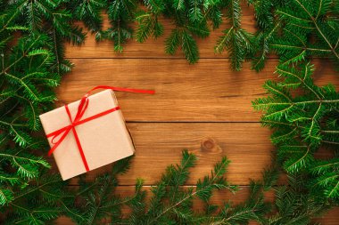 Kırmızı kurdeleli küçük bir hediye kutusu ahşap bir masada oturur. Yeşil çam dalları kutunun etrafını sararak doğal bir sınır oluşturuyor. Bu sahne tatil temaları ve dekorasyonları için uygundur..