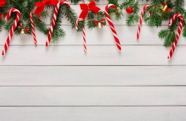 Kırmızı ve beyaz çizgili şeker kamışları yeşil dallar ve kırmızı fiyonklar arasında asılı. Dekorun bir parçası küçük çanlar. Bu ekran beyaz ahşap bir yüzeyde duruyor. Bu da şenlikli bir ortam olduğunu gösteriyor..