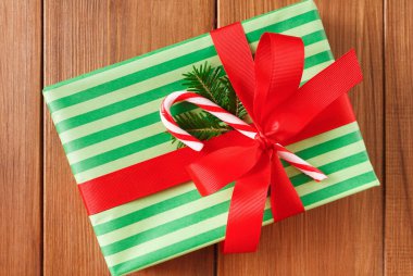 Yeşil ve kırmızı çizgili bir hediye dikkatlice paketlenmiştir. Bir şeker çubuğu ve küçük yeşil bir bitkiyle süslenmiştir. Arka plan, sahneye sıcaklık katan ahşap bir yüzey..