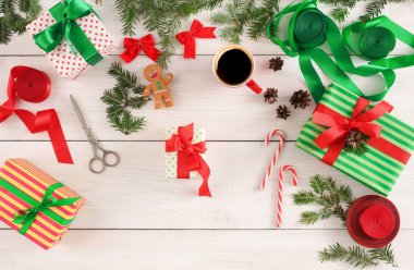 Beyaz ahşap bir masada farklı paketlenmiş hediyeler düzenlenir. Çam dalları, zencefilli kurabiye figürü, kurdeleler, makas ve bir fincan kahve var..