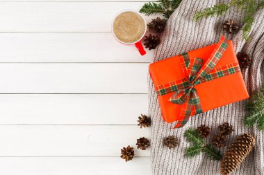 Tatil eşyaları arasında bir paket hediye, bir kurdele, bir fincan sıcak kahve ve yumuşak bir battaniye üzerinde çam kozalakları yer alıyor. Yeşil çam dalları, ahşap bir yüzey üzerinde olay yerinin çevresini sarar..