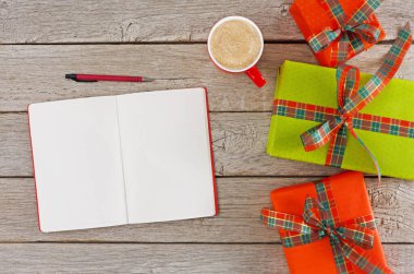 Ahşap bir masa boş bir defter, kırmızı bir kalem, bir fincan kahve ve canlı renklerde üç hediye içerir. Bu ayar, özel bir zamanda yazma ve kutlama için davet edilmektedir.