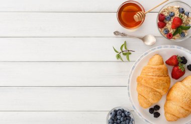 İki kruvasan, böğürtlen dolu kaselerin yanında bir tabakta dinleniyor. Beyaz bir masada bir bardak bal ve bir kase meyveli yulaf ezmesi mevcuttur. Bu sahne bir sabah yemeği düzenini gösteriyor..