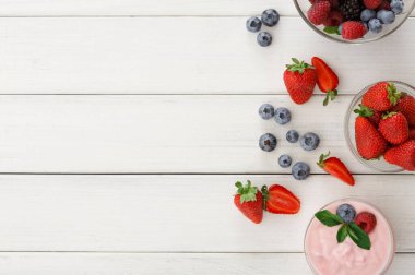 Çeşitli konteynırlarda ahşap bir masada çilek, yabanmersini ve böğürtlen bulunur. Renkli meyvenin yanında bir kase yoğurt duruyor. Düzenleme basit bir aperatif ya da tatlı seçeneği gösteriyor..