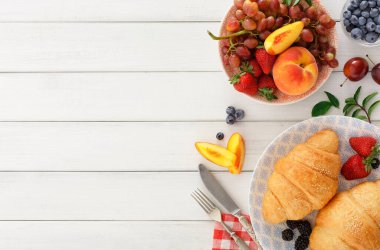 Bir masa iki kruvasan ve karışık meyvelerle hazırlandı. Meyveler arasında üzüm, çilek, şeftali ve yabanmersini yer alıyor. Yakınlarda bir bıçak ve peçete var..