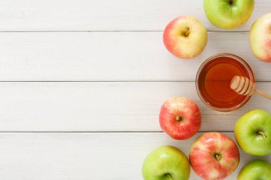 Farklı renklerde taze elmalar bir kavanoz balı ahşap bir masanın üzerinde bir kepçeyle sarıyor. Sahne parlak ve sergilenen doğal bir gıda düzenlemesi genellikle aperatifler veya tatlılar için kullanılır.