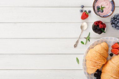 Kruvasanlar bir tabak taze böğürtlenin yanında dinlenir. Yakınlarda bir kaşık yatıyor ve yoğurt olay yerine renk katıyor. Bu kahvaltı masası basit bir sabahı davet ediyor..