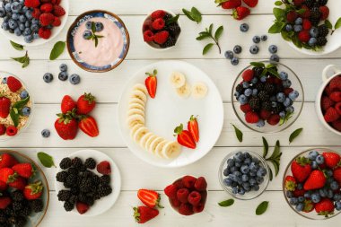 Çeşitli meyvelerle dolu bir kase ve tabak bir masanın üzerine konur. Çilek, yabanmersini, böğürtlen ve muz mevcuttur. Yoğurt kasede..