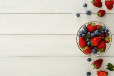 Taze çilekler ve yabanmersinleri temiz bir kasede düzenlenir. Bazı böğürtlenler hafif bir ahşap masaya saçılmıştır. Ayarlar temiz ve basit bir besin gösterisi gösteriyor..