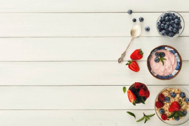 Yoğurt, böğürtlen ve granola dolu farklı kaseler ahşap bir yüzeye dizilir. Taze çilekler ve yabanmersinleri yakınlarda. Bu sahne kahvaltı için mükemmel..