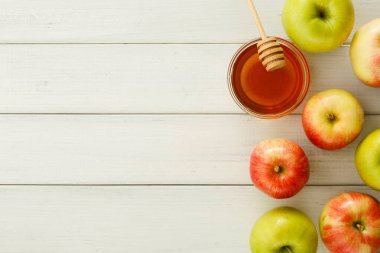 Kırmızı ve yeşil elmalar, açık ahşap bir masada bal kavanozunun etrafını sarar. Sahne, meyvenin doğal renklerini ve balın tatlılığını yakalar. Bu düzen bir tazelik hissi uyandırıyor.
