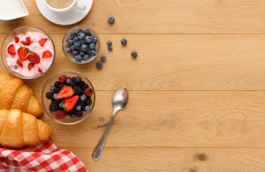 Kahvaltı malzemelerinin bir koleksiyonu ahşap bir masada sergilenir. Meyveli yoğurt, bir fincan kahve ve kruvasan var. Ayarlar renkleri ve dokuları gösterir.