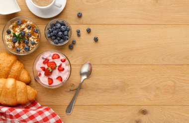 Kahvaltı tabakları ahşap bir masada oturur. Çilekli yoğurt, meyveli tahıl gevreği ve kruvasan var. Yakınlarda bir fincan kahve var..
