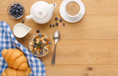 Kahvaltı malzemelerinin bir koleksiyonu ahşap bir masada düzenlenir. Çaydanlık bir fincan kahvenin yanında oturur..