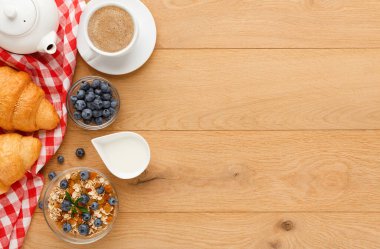 Kahvaltı malzemelerinin bir koleksiyonu ahşap bir masada düzenlenir. Kruvasan, bir fincan kahve, tahıllı yoğurt ve bir kase taze yaban mersini var. Sahne basit ve davetkar..