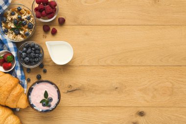 Kahvaltı malzemelerinin bir koleksiyonu ahşap bir masanın üzerine yayılır. Kaselerde yoğurt, yabanmersini ve ahududu var. Bir kruvasan ve tahıl da dahil..