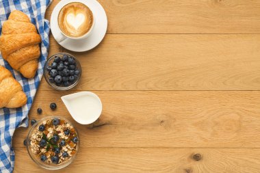 Kahvaltı malzemelerinin bir koleksiyonu ahşap bir masada düzenlenir. Kruvasan, köpük sanatı, yabanmersini, yoğurt ve küçük bir kap süt ile bir fincan kahve hoş bir sabah sahnesi yaratır..