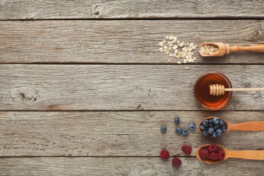 Kahvaltı için farklı malzemeler içeren bir tahta kaşık seti; yulaf, bal, yabanmersini ve ahududu içerir. Arkaplan, temaya eklenen rustik ahşap bir tablo gösterir.