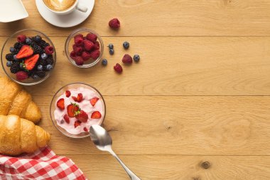 Kahvaltı malzemelerinin bir koleksiyonu ahşap bir masada sergilenir. Kaselerde yoğurt ve taze böğürtlen var. Bir kruvasan bir fincan kahvenin yanına oturur ve bir sabah yemeği sahnesi yaratır..