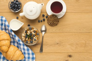 Bir kolaj ahşap bir masanın üzerinde çeşitli kahvaltı öğelerini gösterir. Çay, kruvasan, yaban mersini ve tahıllı yoğurt var. Bu düzen bir sabah yemeği deneyimini davet ediyor..