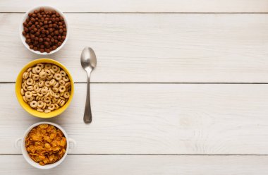 Üç kase kahvaltı gevreği ahşap bir masaya konur. Kâselerin yanında bir kaşık var. Gevrekler yuvarlak parçalar, gevrek gevrekler ve çikolata toplarını içeriyor..