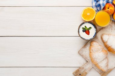 Kahvaltı malzemeleri ahşap bir yüzeye dizilir. Kruvasan, meyve, yoğurt ve bir bardak meyve suyu var. Sahne, çeşitli yiyeceklerle basit ve davetkar bir sabah yemeği düzenini gösteriyor..