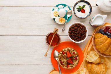 Kahvaltı yiyeceklerinin bir koleksiyonu ahşap bir masada sergilenir. Tabaklar ve kaseler mısır gevreği, yoğurt, böğürtlen, yumurta ve bal içerir. Kruvasanlar ve diğer malzemeler yayılıma eklenir..