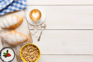 Kahvaltı malzemeleri ahşap bir masada sergilenir. Kruvasanlar tahtaya konur. Kalp sanatı olan bir kahve fincanı sarı bir kase mısır gevreği ve meyveli küçük bir yoğurdun yanında oturur..