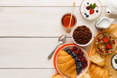 Farklı kahvaltılık yiyecekler ahşap bir masaya konur. Sahnede hamur işleri, meyveler, tahıllar ve içecekler yer alıyor. Parlak renkler ve çeşitli dokular yemek için davetkar bir görünüm yaratır..