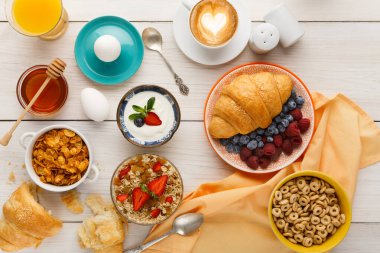 Farklı kahvaltı öğeleri ahşap bir masada sergilenir. Meyve, yoğurt, tahıl, kahve ve yumurta var. Tatlım, kruvasan ve içecekler yemek seçeneklerini tamamlar..