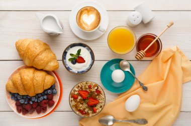 Kahvaltı malzemelerinin bir koleksiyonu ahşap bir masada düzenlenir. Kruvasan, yoğurt, meyve, bal, yumurta ve içecekler mevcut. Bu set farklı kahvaltı seçeneklerini vurgular.