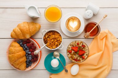 Kahvaltı malzemelerinden oluşan bir kolaj ahşap bir masada düzenlenir. Kruvasan, meyve, mısır gevreği, yoğurt, içecek ve karıştırmak için aletleri olan bal var. Tüm ögeler düzgünce görüntülenir.