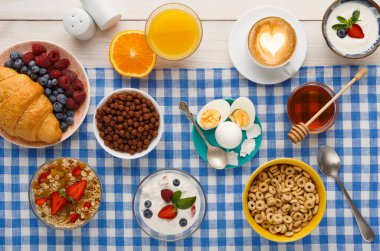 Kahvaltı malzemelerinin bir koleksiyonu kareli bir masa örtüsüne yerleştirilir. Kaselerde meyve, mısır gevreği ve yoğurt var. İçecekler arasında portakal suyu ve kahve yer alıyor.