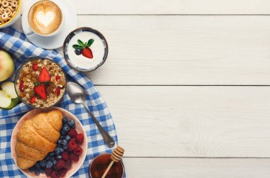 Kahvaltılık eşyalar kareli bir kumaşın üzerinde sergilenir. Meyve, yoğurt, tahıl gevreği, kahve ve kruvasan var. Sahne, çeşitli lezzetlerde sıradan bir sabah yemeği gösteriyor..