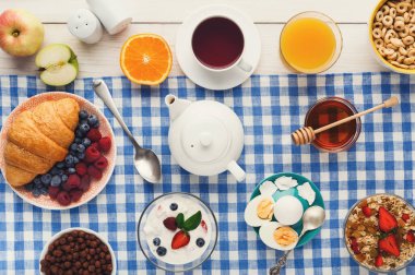 Kahvaltı yiyeceklerinin bir koleksiyonu masanın üzerinde düzenlenir. Meyve, mısır gevreği, yoğurt, çay, meyve suyu ve hamur işleri var. Bu ayar bir sabah yemeği için çeşitli seçenekleri gösterir.