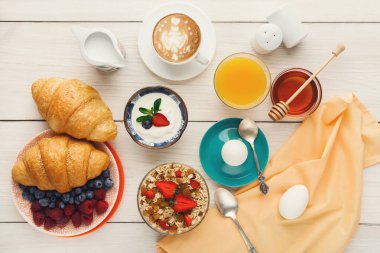 Bir koleksiyonda hafif ahşap bir masada çeşitli kahvaltı öğeleri yer alır. Kruvasan, böğürtlenli yoğurt, yumurta, bal ve içecek var. Düzenleme çeşitli tatlar gösteriyor..