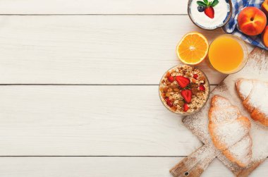 Bir kolaj ahşap bir masanın üzerinde çeşitli kahvaltı gıdalarını gösterir. Kruvasan, meyveli yoğurt, bir kase granola ve meyve suyunun yanında portakal dilimleri var..