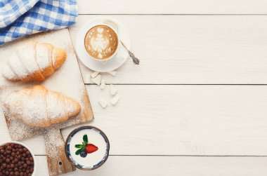 Bir kahvaltı öğeleri koleksiyonu ahşap bir yüzey üzerinde sergilenir. Pudralı iki kruvasan, bir fincan kahve ve üstünde çilek ve böğürtlen olan bir kase yoğurt var..